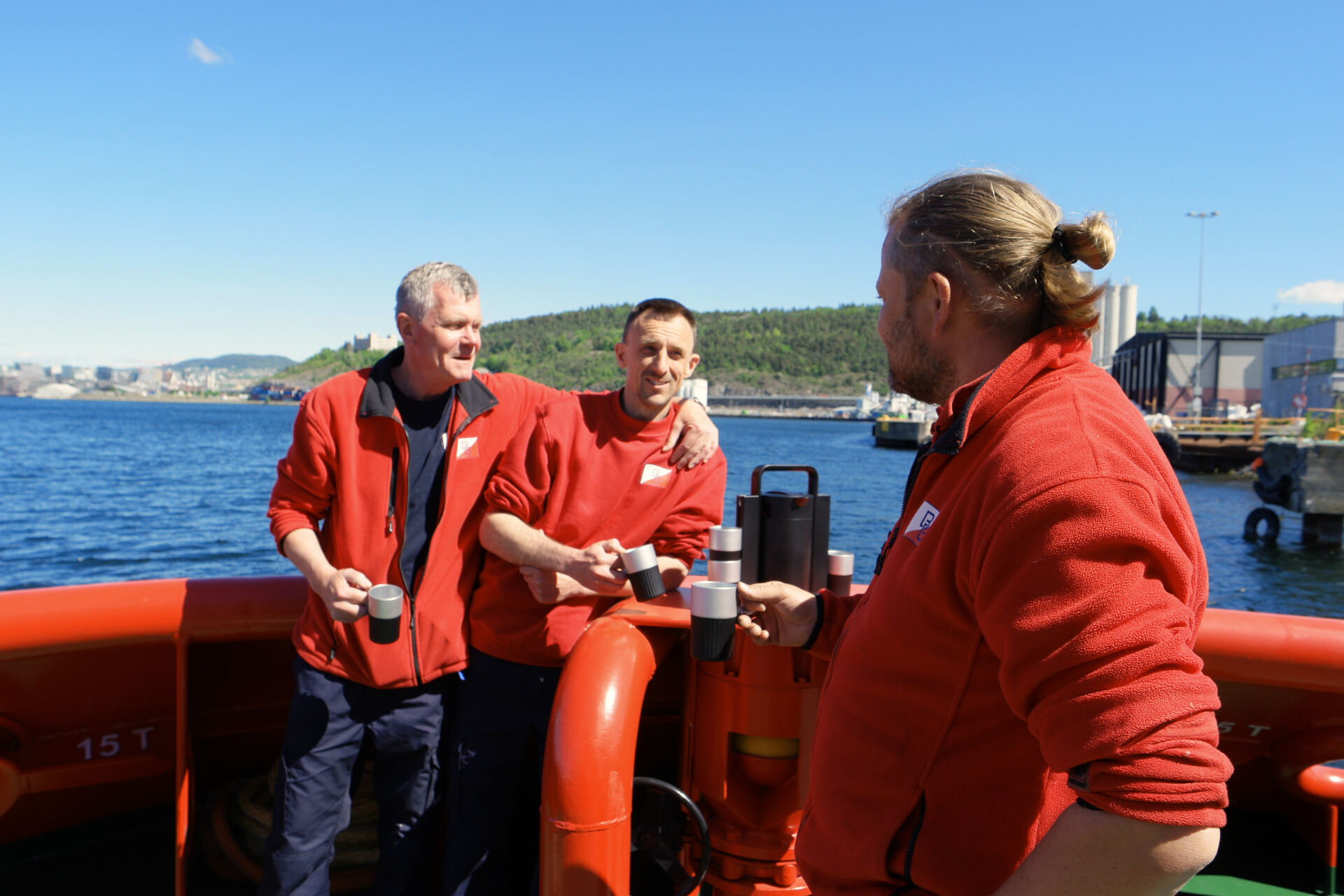 Tre sjømenn i røde jakker står på dekk og drikker fra hver sin kopp ved siden av kaffesystemet. Den ene mannen har armen rundt en kollega, og de smiler i sola med havn og skogkledde åser i bakgrunnen.